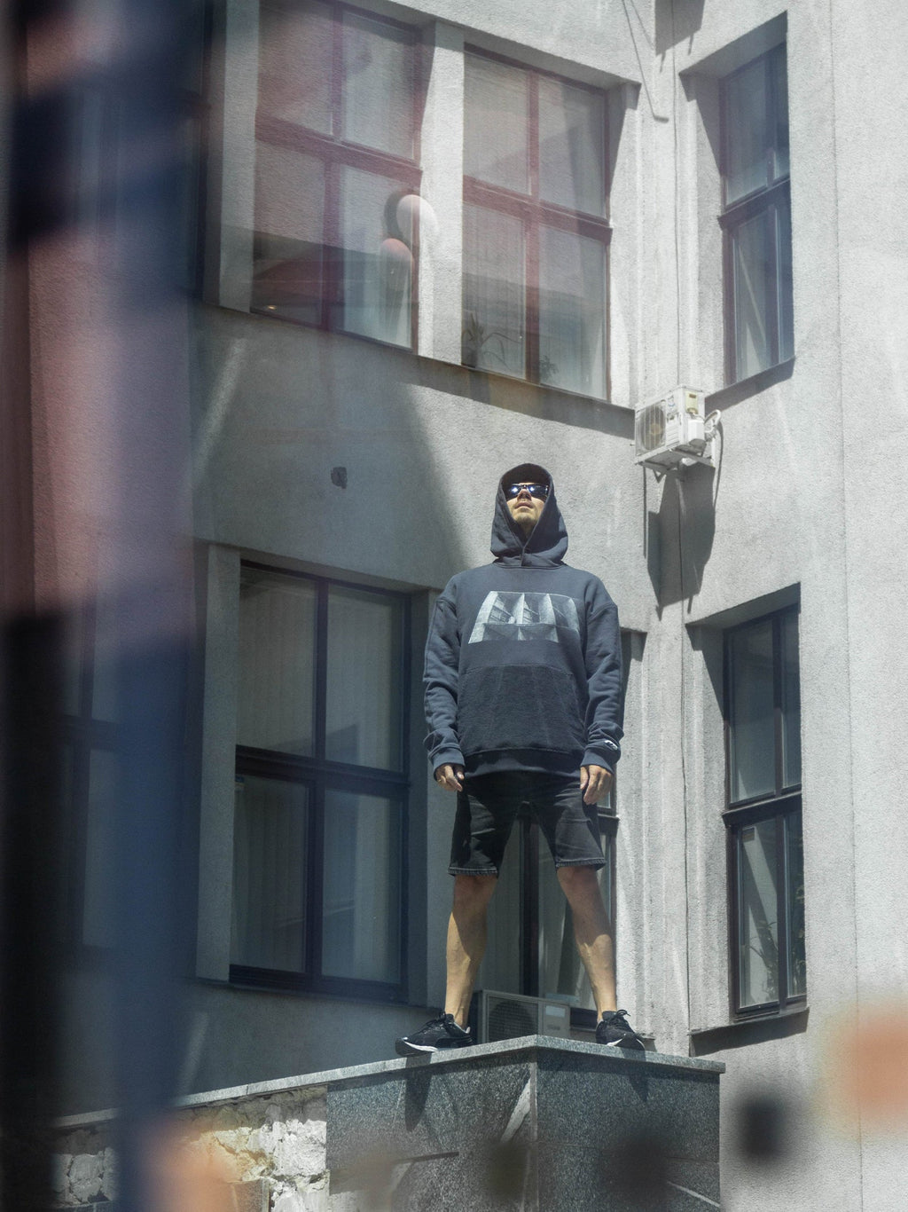 Person wearing a dark hoodie with a brutalist print in front of a building.