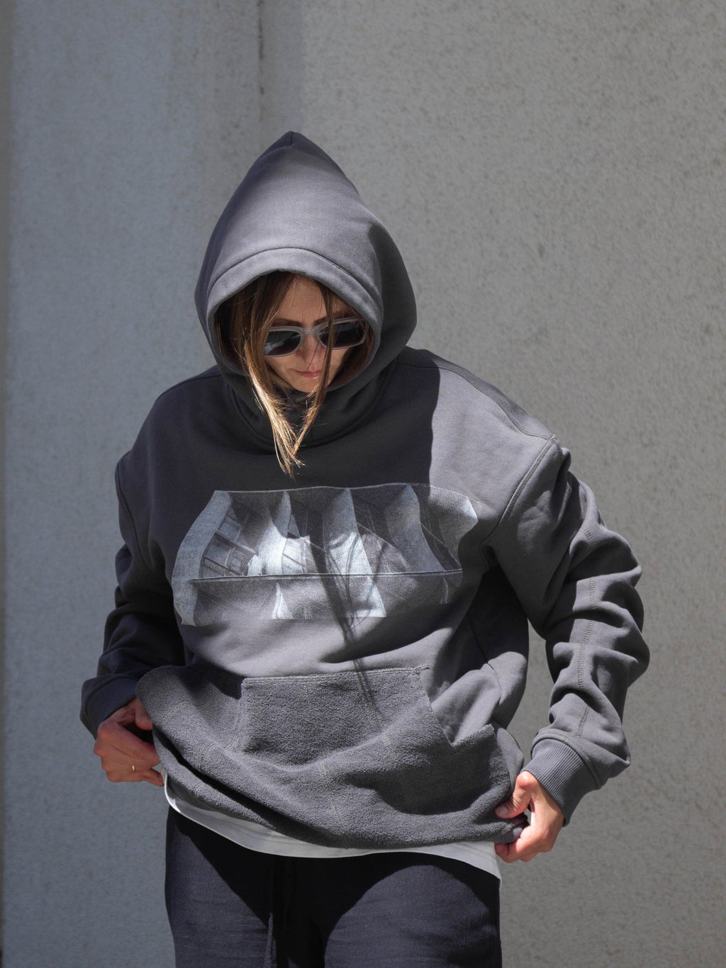 A person wearing an oversized dark gray hoodie with a brutalist print on the front, styled with dark pants and sunglasses, standing against a textured gray wall.